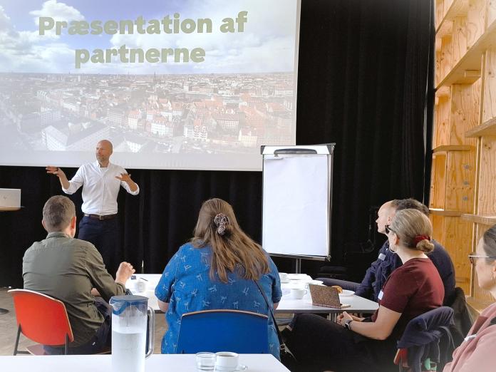 Foredragsholder foran forsamling med teksten præsentation af partnerne på overhead